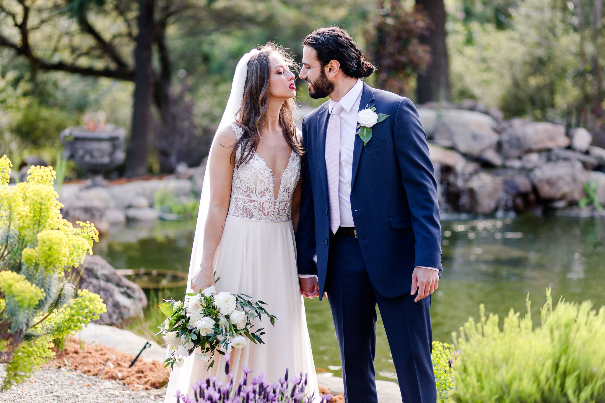 Bride and groom leaning in to kiss, portrait in front of pond with white floral bouquet and flower scenery