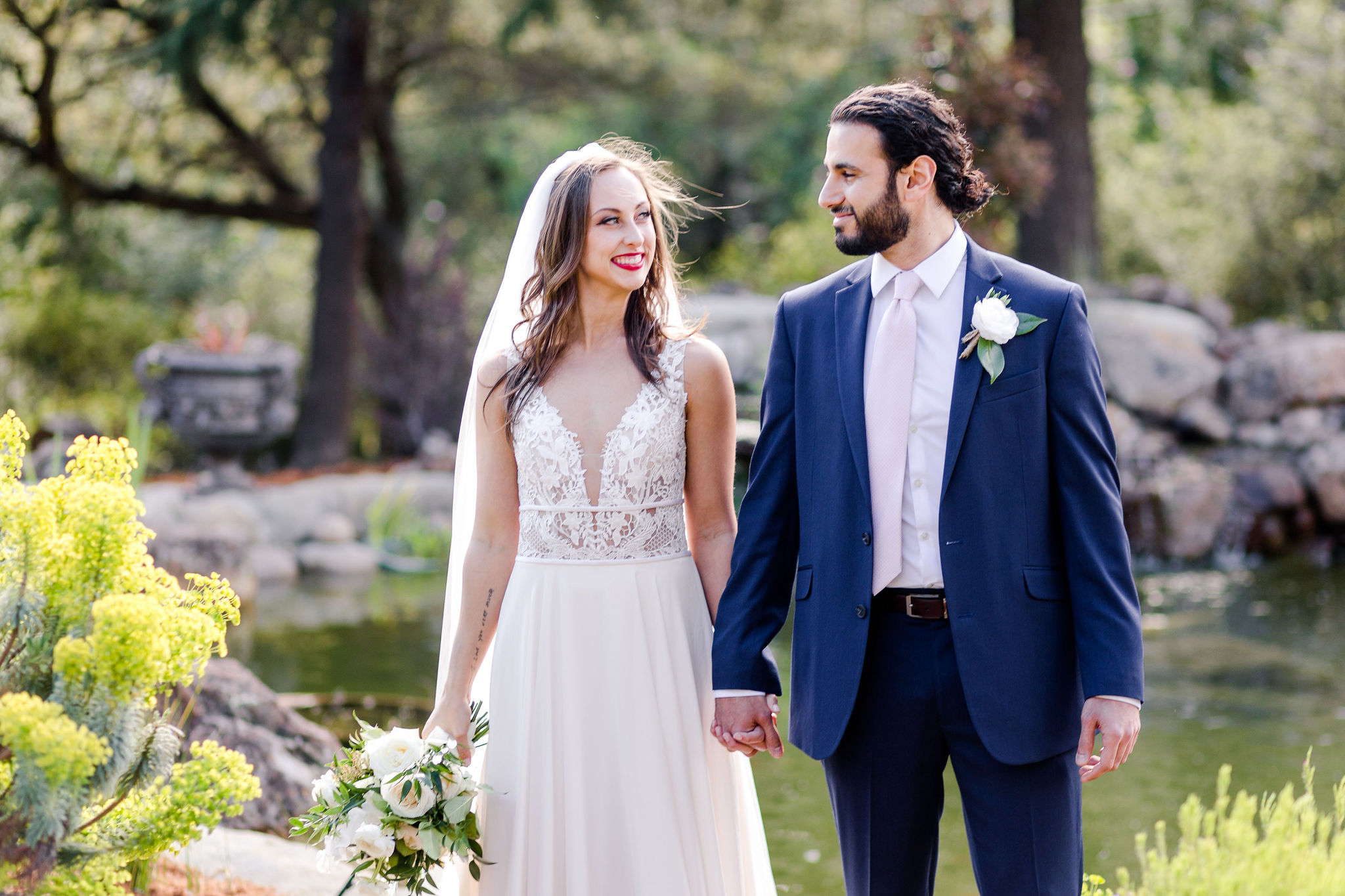 Bride and groom portrait in front of pond with white floral bouquet and flower scenery
