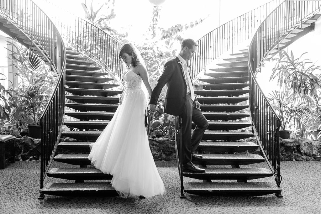 Black and white photo of bride and groom praying together back-to-back before the ceremony begins. Double staircase in the lobby of Ceres Seventh-day Adventist Church. Shot by Amber Rivas Photography.