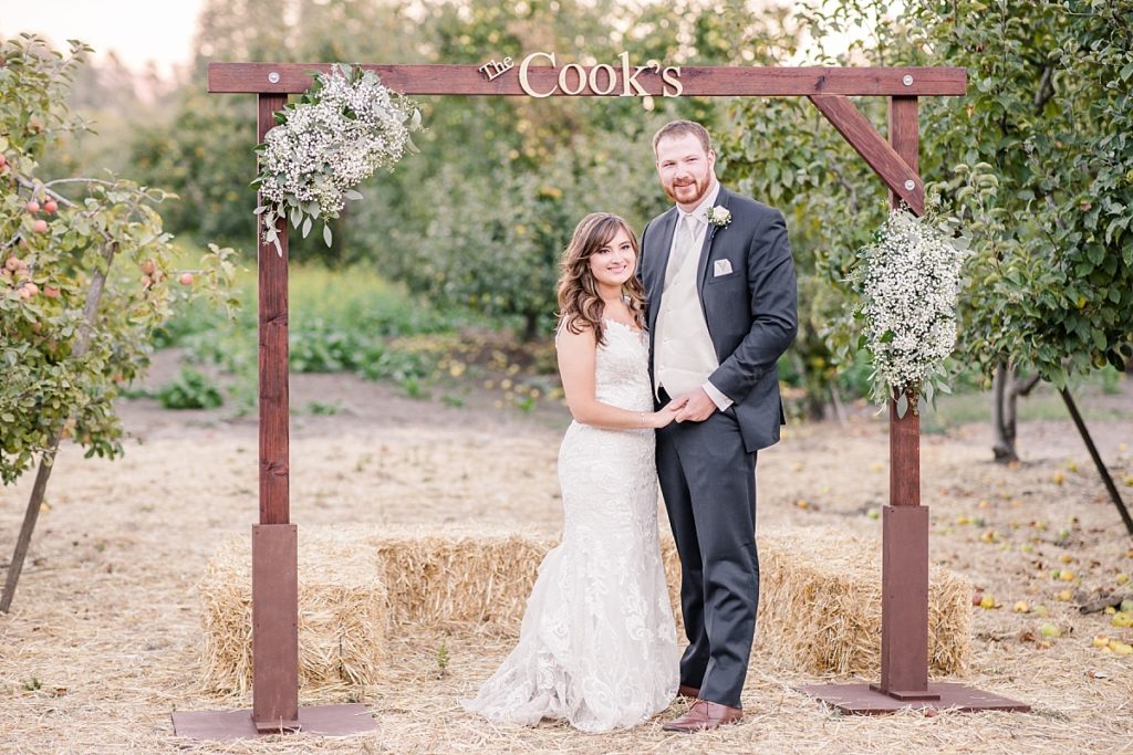 Bride and groom under their wedding arch in the apple orchard at The Orchard in Watsonville, CA. Fall colors in the background, apple trees everywhere.