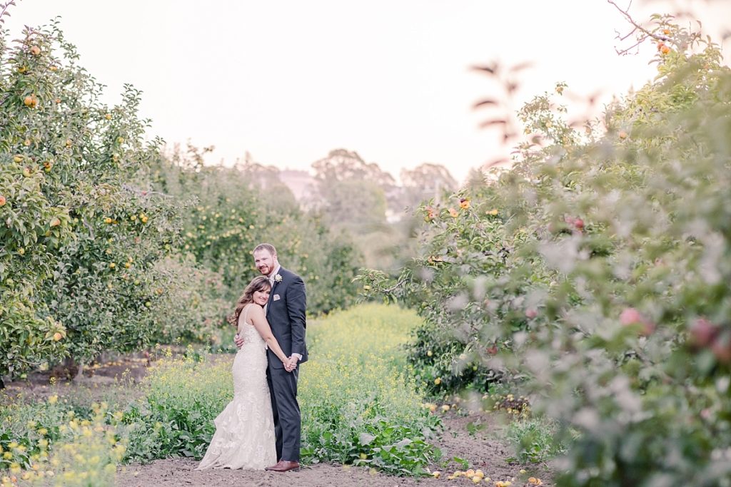 Bride and groom hug and smile in the apple orchard at The Orchard in Watsonville, CA. Shot by Amber Rivas Photography.
