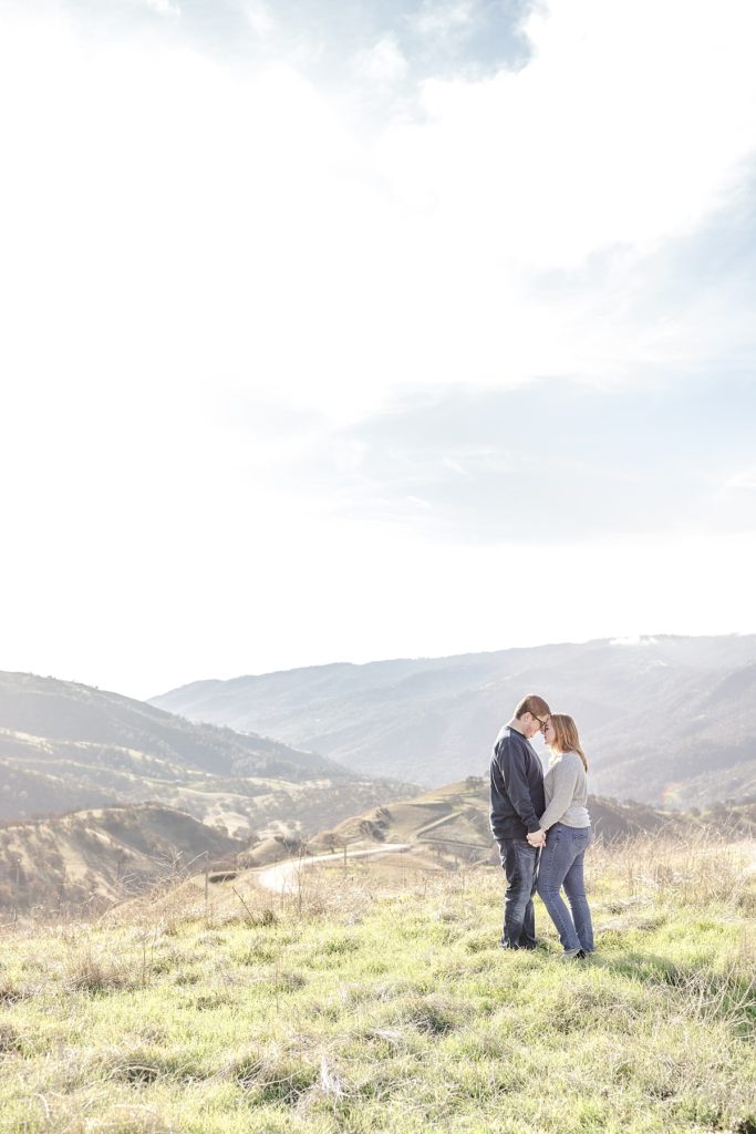 Del Valle engagement photos by Livermore photographer. Couple sunlit standing at Top of the Hill overlooking hills.