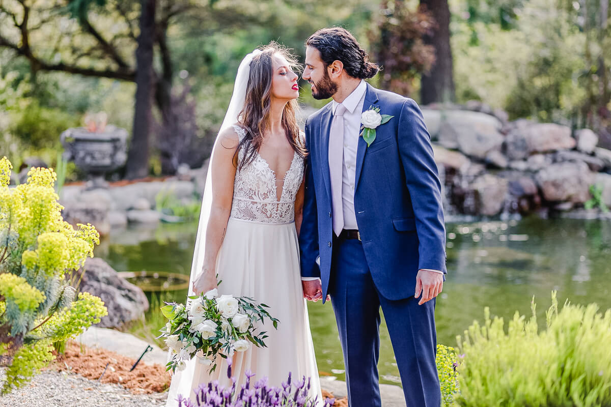 Bride and groom leaning in to kiss, portrait in front of pond with white floral bouquet and flower scenery