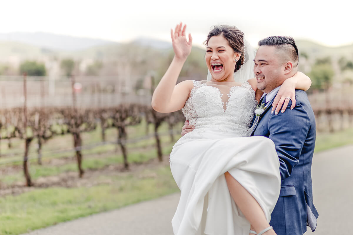 Groom holds bride who laughs and waves in the vineyard at Palm Event Center