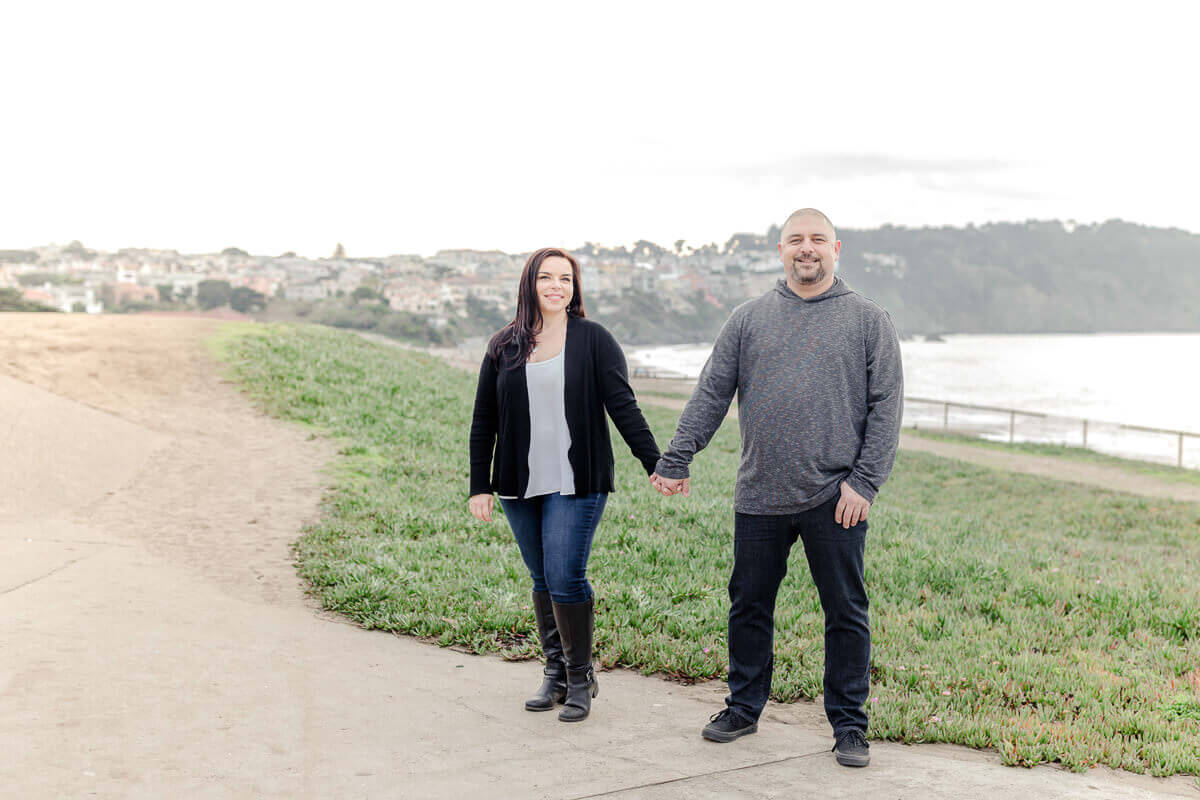 Man and woman smile at the camera holding hands, standing on a path overlooking Baker Beach in San Francisco.