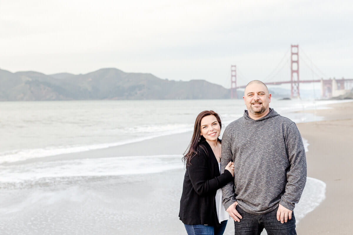 Man and woman smile at the camera in front of the Golden Gate Bridge at Baker Beach in SF.